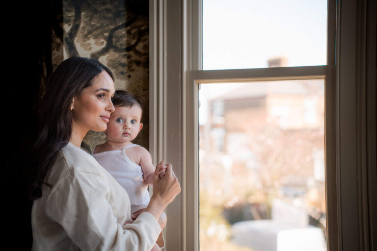 Mother holding baby while looking out a window during a natural light motherhood photography session in Munich.